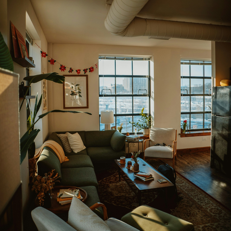 Cozy modern apartment living room with large windows, sectional sofa, and warm natural lighting.